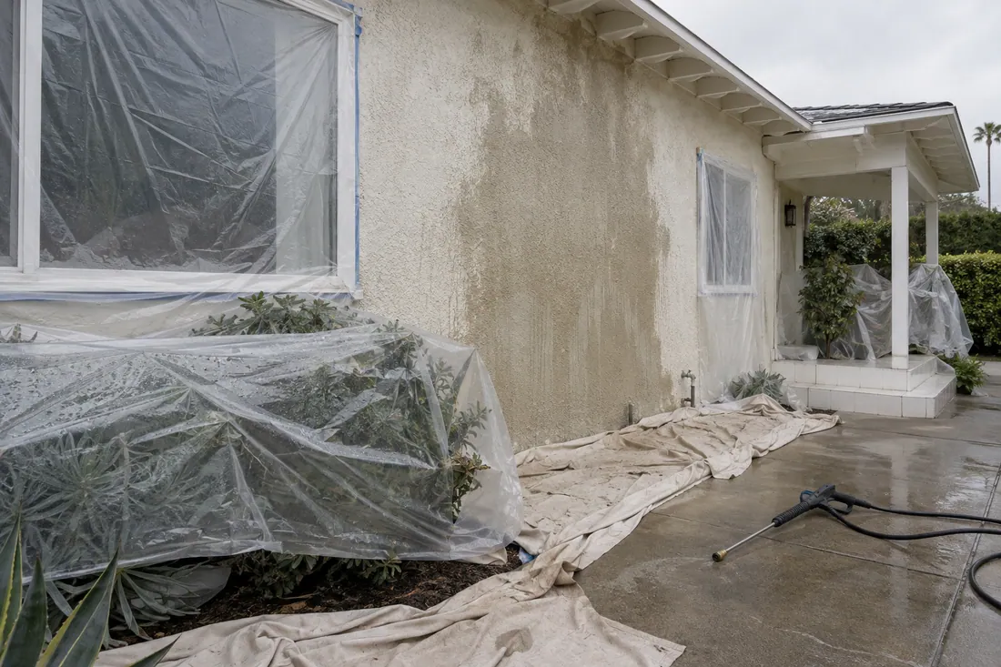 Stucco wall wet from low-pressure washing before exterior painting, with plastic over the plants