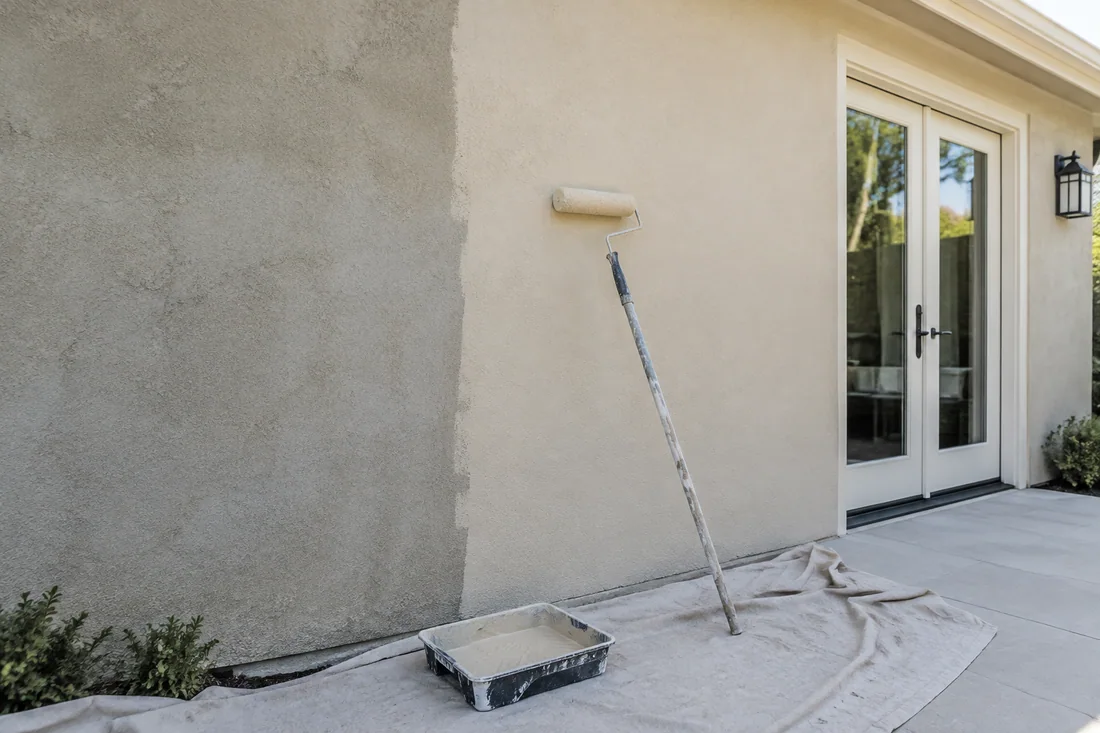 Half-painted stucco wall with a roller and tray, showing fresh coating beside the faded old color