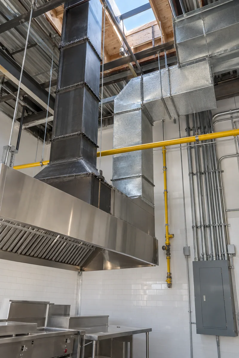 Open ceiling over a commercial kitchen showing a grease duct, a makeup air duct, gas piping and conduit